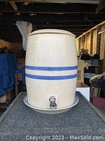 Front view of beige stoneware crock with blue double stripe and metal spigot on the lower front. Crock is placed on matching stoneware plate.