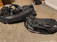 Two black genuine leather duffle bags on a carpet, showing the relative size and handles of both bags.
