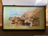 Framed artwork displayed against plywood background, full view of frame, glass front reflecting light