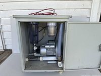 Wide shot of the water suction pump installed inside a gray metal cabinet with hinged door open. Shows motor, tubing, and filter systems.