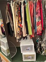 Closet view showing hanging ladies clothing including tops and pants in multiple colors and patterns, white plastic storage drawers below