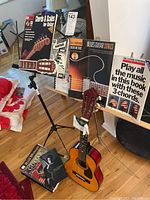 Photo showing the junior guitar placed on floor near two music stands holding various guitar sheet music and song books.