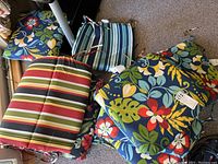 Overview photo showing eight outdoor chair cushions and one large cushion arranged on carpeted floor. Includes floral and striped patterns.
