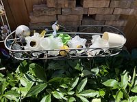 Metal scrollwork tray stand holding frosted glass lamp shades and a white plastic pot, with a metal hummingbird ornament