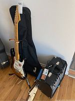 Full overview: Squier Stratocaster on stand, two soft cases behind, Fender Frontman amp with cables and Boss tuner on top