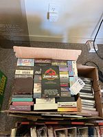 Box filled with assorted VHS tapes and DVDs, titles include Jurassic Park, My Fair Lady, Elvis Presley, Clint Eastwood movies, some in slipcases, some unopened.