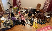 Wide angle photo showing multiple pairs of women's shoes and boots arranged on wooden floor with some boots standing upright along the wall.