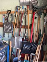 Wide shot of multiple yard tools leaning against a wall including snow shovels, rakes, long-handled hand saws, and other yard implements.