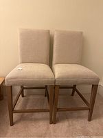 Two cream-colored upholstered wooden chairs side by side showing full front view and legs.