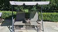 Full front view of the double seat glider patio chair showing the beige fabric seating and canopy, metal frame, and center glass table.