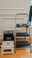 Photo of three step stools: one tall metal frame Cosco 3-step stool with black steps and two smaller plastic folding stools in white and black.