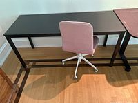 Table with black metal legs and pink office chair from a side angle showing the full table and chair setup on a wooden floor with wall baseboard.