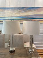 Pair of table lamps with clear glass cylindrical bases and white drum shades placed on a wooden surface, with a painting and beige upholstered furniture in the background.