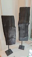 Two black painted wood sculptures on metal stands displayed on a window sill, showing relative height difference and details.