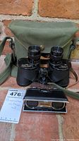 Large binoculars placed in front of an olive green carrying case with a strap, set on a brick surface with auction tag visible.