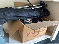 Cardboard box containing assorted camera accessories and a black carrying bag with tripod inside