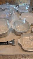 Multiple CorningWare casserole bowls in white with blue cornflower design, each with a glass lid, along with a black-handled plastic utensil on a cloth.
