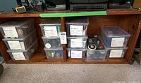 Clear plastic storage totes stacked inside wooden shelving. Each tote is labeled with contents such as wood screws, nails, and machine screws. Visible small containers and tins inside the totes show assorted fasteners.