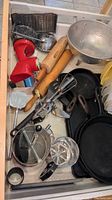 Drawer with assorted kitchen utensils including cast iron fry pans, rolling pins, hand mixers, metal graters and strainers, and red food grinder.