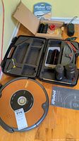 Full overview of the orange iRobot Roomba vacuum cleaner along with accessories arranged: brushes, battery, charger, manuals in box and foam carrying case
