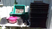 Photo of multiple stacked black soil screener trays, various garden pots including pink colander pot, black ridged pots, green garden edging, and a small black funnel on table.