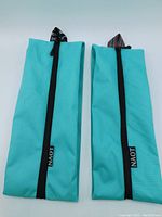 Two turquoise NAOT travel shoe bags with black zipper closures and patterned hanging tags laid flat on white background.