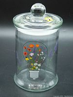 Clear glass cylindrical apothecary jar with a removable glass lid featuring a round handle. One side decorated with a colorful floral light bulb graphic and purple flower accents.