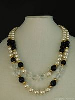 Frontal view of vintage double strand necklace showing alternating large cream faux pearls, clusters of black beads, clear faceted oval beads and gold bead accents on display stand.