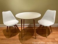 Photo showing the full set: one white round dining table with four wooden legs and two white chairs with wooden legs, placed on wood flooring against a beige wall.