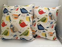 Pair of square decorative pillows printed with multiple colorful birds, standing upright.