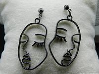 Full view of both Picasso inspired face drop earrings lying flat on a white cloth support. Showcases the black metal wire design of abstract faces hanging from spherical metal studs.
