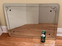 Full front view of rectangular mirror leaning against wall, showing the etched corner details and beveled edges, with two soda cans for scale