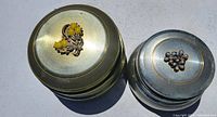 Top view of two round metal powder trinket music boxes with floral ornaments on lid, one brass-colored with yellow beads and one silver-colored with purple beads.