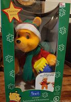Front view of animated Winnie the Pooh Christmas ornament inside original box showing the character in a Santa hat and scarf holding a honey pot.