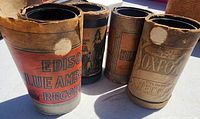 Four Edison Blue Amberol cylinder record containers showing original paper labels, some torn and separating, with visible wear and discoloration.