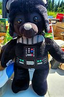 Black plush teddy bear with Darth Vader costume, including helmet and printed control panel, sitting on a white and blue surface outdoors with background of trees and boxes.