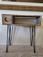 Front view of handmade wooden side table with black metal hairpin legs, small cabinet on left with moon detail, open shelf on right.