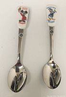 Front view of two spoons showing Mickey Mouse and Donald Duck character heads on white tops with metal spoon bowls.