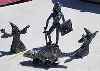 Photo showing a female warrior figurine holding sword and shield, two wizard-like figures with pointed hats, and a dinosaur figurine on a light surface in bright sunlight.