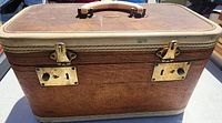 Vintage brown faux leather train case with cream trim and handle. Exterior shows wear and patina. Gold-tone metal locks on front.