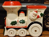 Front and right side view showing Santa figure and holly decoration on the ceramic Christmas train bank with red roof and white body.