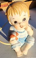 Close-up image of ceramic child figurine night light, showing facial features and details, seated and holding a ball, with beige and blue colors.