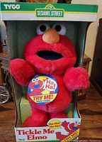 Photo of the front of the original box showing red Tickle Me Elmo plush toy inside with promotional tag to try the giggling and shaking feature, Tyco and Sesame Street branding visible on box.