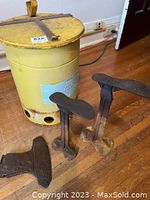 Photo showing yellow tin waste bucket, two cobbler shoe stands, and iron axe head on wooden floor