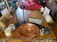 Full view of lot items on wooden table including black cast metal candle holders, hammered copper bowl, stone vase, stone stands, ceramic candle holder and glass candle.