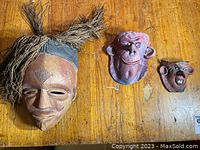 Three masks displayed on wood surface: one South American tribal ceramic mask with straw hair, two smaller ceramic ELF face masks side by side.