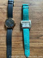 Two designer watches laid flat on a wooden surface. Left watch has a round black dial with black leather band and gold-tone accents, right watch has a rectangular white dial and turquoise textured leather band.