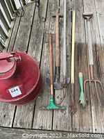 Photo shows large red vintage farm milk jug and an assortment of garden tools with wooden handles laid out on a wooden deck