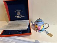 Miniature enamel teapot with colorful artwork of cats and buildings with red textured box with blue satin interior labeled 'Numbered Edition Charlotte di Vita MBE Collections Limited'.