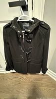 Front view of dark navy wool pea coat hanging on white hanger showing zipper and toggle front closure with two pockets and shoulder button details.